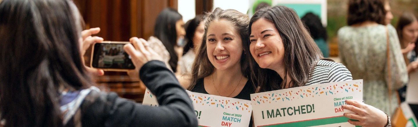 Two women smiling and holding signs that say I Matched while a third person takes their picture
