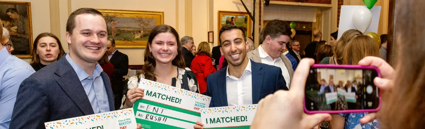 Three medical students smiling and holding signs that say I Matched, with hands holding a phone to take a photo seen in the foreground 